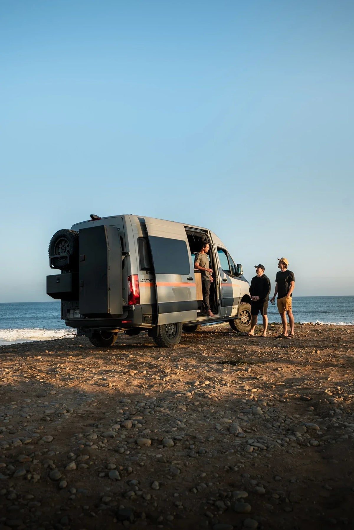 Three people talking beside a van parked on a beach at sunset